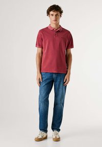 Camiseta polo roja para hombre, de manga corta, tejido de algodón, con cuello y detalle de logo. Combinada con vaqueros azul y zapatillas blancas.