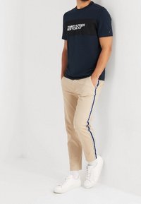 Mand iført en navy Tommy Hilfiger T-shirt, beige bukser med blå-hvide side striber, hvide sokker og hvide sneakers, stående med hænderne i lommerne.