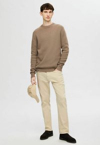 Maglione a maglia marrone chiaro, pantaloni di cotone beige, tiene in mano un berretto beige, mocassini neri, in piedi contro uno sfondo semplice.