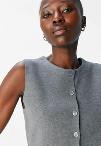 Gilet sans manches gris, avec encolure ronde, fermeture à cinq boutons et texture côtelée. Comprend des ouvertures sur les côtés pour une meilleure ventilation.