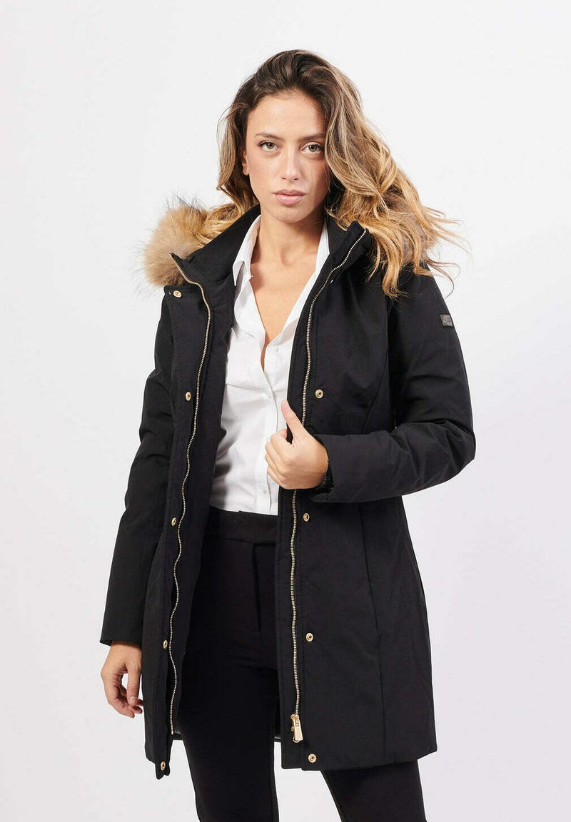 Donna con un cappotto invernale nero con cappuccio di pelliccia, aperto per rivelare una camicetta bianca, in piedi contro uno sfondo chiaro e uniforme.