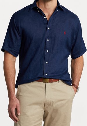 Homme portant une chemise bleu marine à manches courtes boutonnée avec un logo rouge, pantalon beige, ceinture marron et montre-bracelet, main dans la poche.