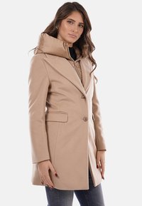 Cappotto color cammello con design strutturato, bottoni doppiopetto e cappuccio trapuntato removibile. Texture liscia con tasche laterali.
