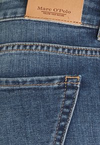 Poche arrière d'un jean en denim bleu avec des coutures marron et une étiquette de marque en cuir beige portant l'inscription « Marc O'Polo Follow Your Nature ».