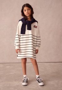 Hvid langærmet kjole med marineblå vandrette striber, med rund hals og et brystpatch. En marineblå kabelstrikket sweater er draperet omkring halsen. Sorte sneakers fuldender looket.