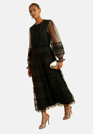 Schwarzes, gestuftes Maxikleid mit gepunktetem Netz, langen durchsichtigen Ärmeln und Rüschendetails. Kombiniert mit einer metallischen Clutch und Riemchen-Heels.