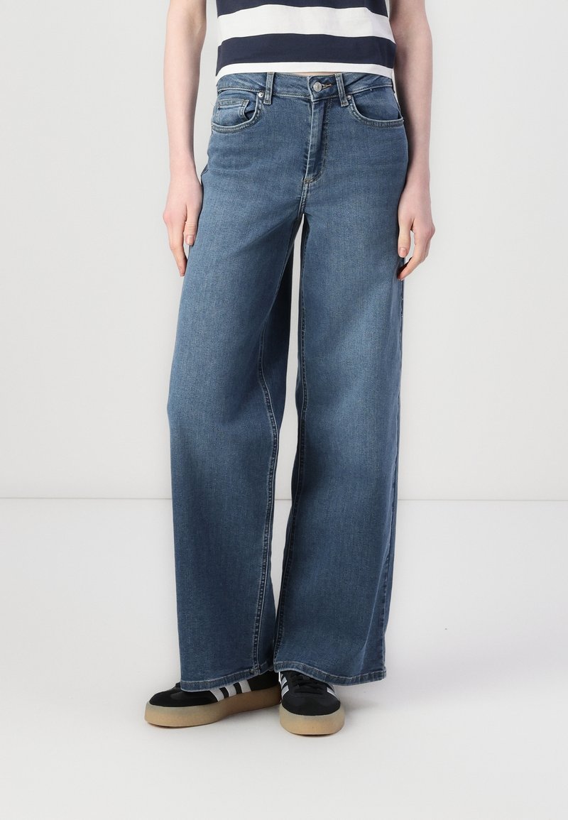 Personne portant un jean large en denim bleu, des baskets noires avec des rayures blanches, et un t-shirt rayé noir et blanc.