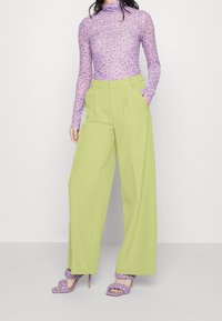 Světlezelené kalhoty s širokými nohavicemi a vysokým pasem, spárované s dlouhým rukávem fialového vzorovaného topu a fialkovými pletenými podpatky.