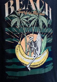 Σκελετός που φοράει καπέλο τρώει μπανάνα καθισμένος πάνω σε μια μεγάλη μπανάνα με φοίνικες και σανίδες σερφ στο παρασκήνιο.