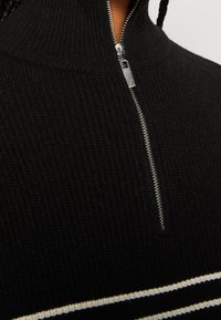 Gros plan d'un pull en tricot côtelé noir avec une fermeture éclair argentée partiellement fermée et un détail à rayures blanches près du bord inférieur.