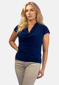 Granatowa bluzka z drapowaną dekoltą, krótkimi rękawami, miękkiego materiału i marszczeniem w talii, zestawiona z beżowymi spodniami.