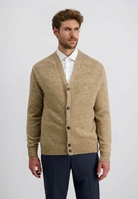 Mann in beigem, geknöpftem Cardigan über weißem Hemd und dunkler Hose, der vor einem hellgrauen, schlichten Hintergrund steht.