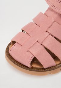 Rosa lädersandal med ett vävt remdesign, platt sula och texturerad trähäl. Velcro-stängning för säker passform.