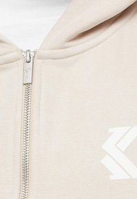 Béžová mikina na zips s strieborným zipsom, bielym logom a textúrovanou tkaninou. Viditeľné sú aj detaily kapucne a vrecka.