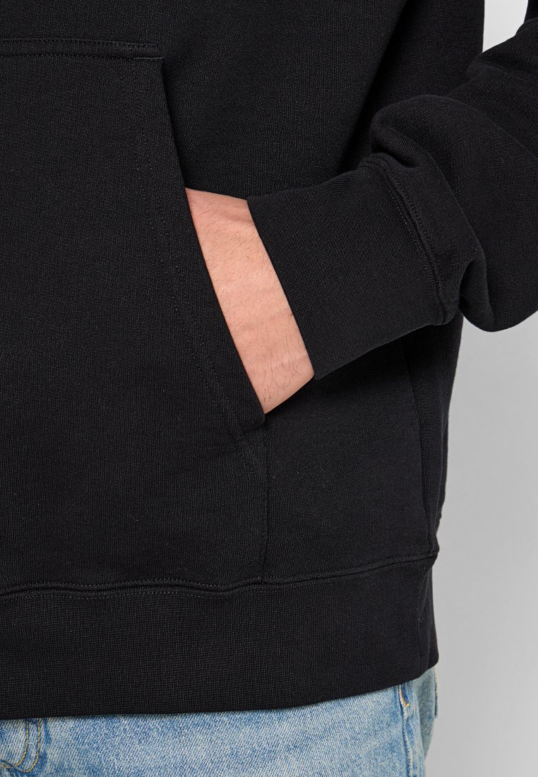 Schwarzer Kapuzenpullover mit einer Fronttasche, gerippten Bündchen und Saum; weicher, strukturierter Stoff; in hellblaue Jeans gesteckt.