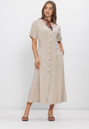 Vrouw met een beige midi-jurk met knopen en korte mouwen, een bordeauxkleurige sjaal met patroon om haar nek en zwarte sandalen met open tenen, staand tegen een effen achtergrond.