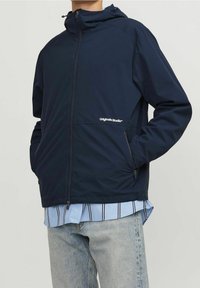 Giacca impermeabile blu navy con cappuccio, dotata di cerniera frontale e tasche laterali. Logo "Originals Studio" in bianco. Tessuto leggero.