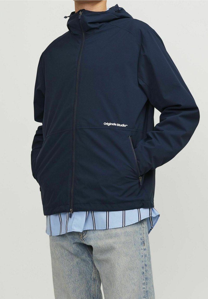 Giacca impermeabile blu navy con cappuccio, dotata di cerniera frontale e tasche laterali. Logo "Originals Studio" in bianco. Tessuto leggero.