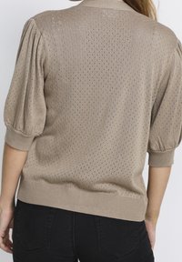Beige stickad tröja med puffärmar, med ett mönstrat textur av små hål och ribbad nederkant. Enkel, avslappnad design.