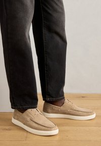 Pantofi slip-on din suede bej cu talpă din cauciuc alb, având cusături decorative și șnururi minime. Purtați cu blugi închisi.