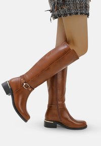 Botas de cuero marrón hasta la rodilla con punta redonda, cierre de cremallera, detalle de hebilla lateral y un tacón bajo apilado. Textura lisa y diseño elegante.