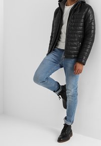 Man som bär en svart vadderad jacka, ljusgrå sweatshirt, ljusblå jeans och svarta snörskor, lutad mot en grå vägg.