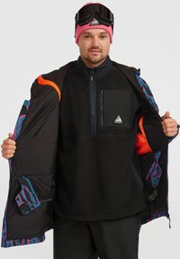 Melns vilnas žakets ar oranžiem akcentiem, rakstainā oderē, ar rāvējslēdzēja krūšu kabatu, un melnas bikses. Valkāts ar rozā beanie un brillēm.