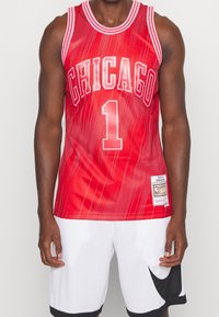 Punainen koripallopaita, jossa on teksti "CHICAGO" ja numero "1". Ominaisuuksiin kuuluu raidallinen kaulus ja teksturoitu pinta. Yhdistetty valkoisten shortsien kanssa, jotka korostavat muotoilua.