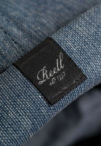 Tejido de mezclilla azul con una etiqueta negra que presenta "Reell" en cursiva blanca y "est. 1993." La textura está ligeramente desgastada con costuras visibles.