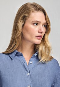 Ljusblå linningblus med krage och knapprad; har vita knappar och en broderad logotyp på bröstet.