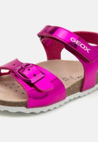 Lapsen vaaleanpunaiset metallinhohtoiset sandaalit, joissa on säädettävä solki ja tarranauha korkkipohjalla. Sandaalien nilkkahihnassa on "GEOX" -brändin logot.