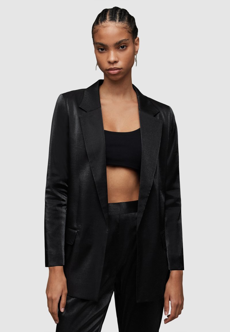 Schwarze Satin-Blazer mit einem Ein-Knopf-Verschluss, spitzem Revers und seitlichen Taschen, getragen über einem schwarzen Cropped-Top. Glatte Textur.