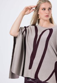 Poncho beige con diseño abstracto en burdeos oscuro, corte holgado, mangas cortas, textura suave, dobladillo que cae por debajo de la cintura, llevado sobre pantalones oscuros.