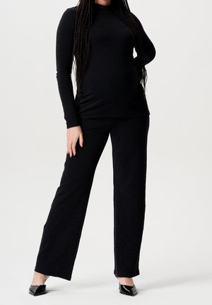 Kvinde iført sort langærmet top og bukser med vide ben, stående med den ene hånd på hoften og sorte spidsede hæle på hvid baggrund.