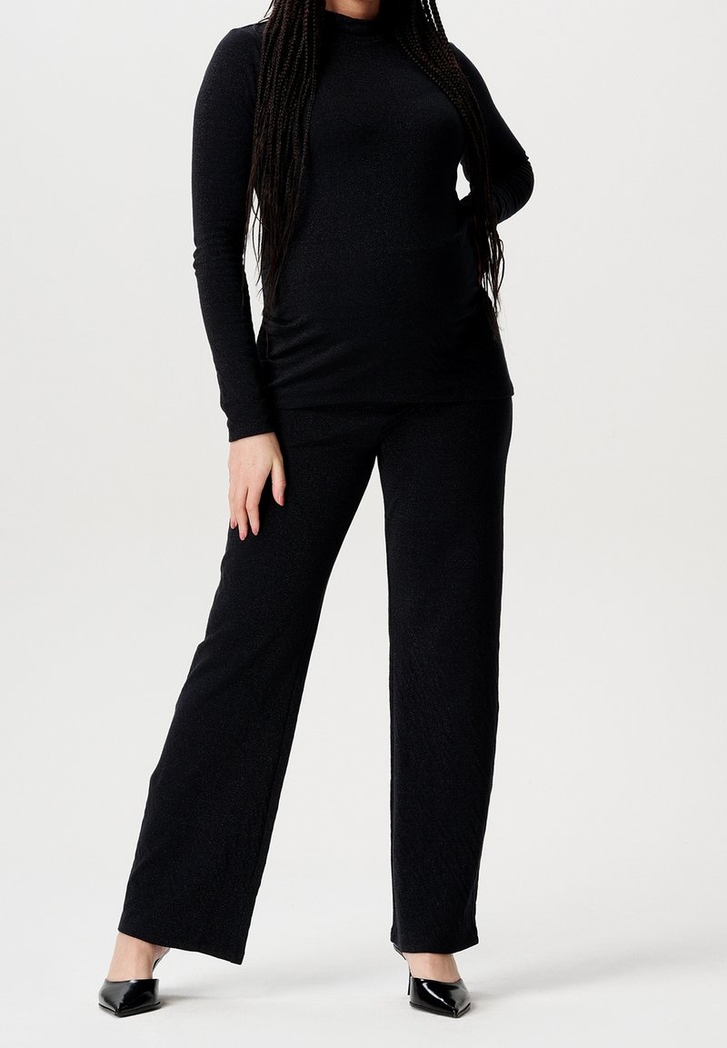 Femme portant un haut noir à manches longues et un pantalon à jambes larges, debout avec une main sur la hanche et des talons noirs pointus sur fond blanc.