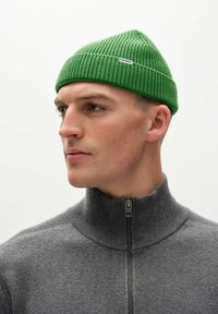 Zeleni rebrasti beanie od pletene tkanine ima uski dizajn s oznakom logotipa. Nosio se uz sivi zip-svećeničar, pokazujući prilagođeni oblik.