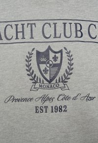 Λογότυπο Yacht Club Monaco με ασπίδα, στέμμα, fleur-de-lis, στεφάνι δάφνης και κείμενο «Provence Alpes Côte d’Azur EST 1982» σε γκρι ύφασμα.
