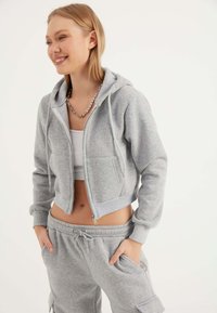 Felpa grigia corta in materiale morbido con chiusura a zip, cappuccio regolabile con coulisse e tasche frontali. Abbinata a pantaloni coordinati.