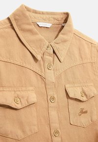 Hiekanvärinen nappipaita teksturoidusta kankaasta, jossa on terävä kaulus, kaksi rintataskua ja vasemmassa taskussa brändätty kirjailu.