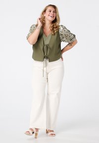 Olijfgroene vest met een gestrikte voorkant, voorzien van versierde pofmouwen. Gecombineerd met crèmekleurige wijde broek en beige hoge sandalen.