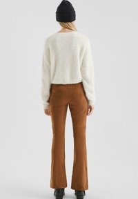 Vrouw in een witte fuzzy trui, bruine suède flared pants, zwarte beanie en zwarte schoenen, staand met haar rug naar de camera tegen een effen achtergrond.
