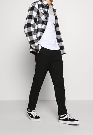 Schwarz-weiß kariertes Flanellhemd über grauem T-Shirt, kombiniert mit schwarzen Hosen und schwarzen Sneakers mit weißen Details und Schnürsenkeln.