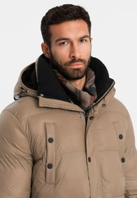 Chaqueta de plumas beige con capucha forrada de forro polar negra, con botones negros, cierre zip en la parte delantera y una bufanda de cuadros marrones alrededor del cuello.