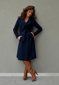 Granatowy dwurzędowy płaszcz wełniany z paskiem, bocznymi kieszeniami i szerokimi klapami, zestawiony z brązowymi wysokimi kozakami na obcasie.