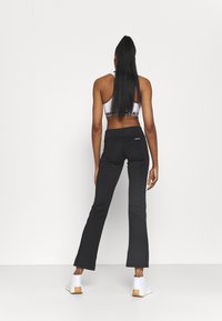 Schwarze, ausgestellte Yogahosen mit hohem Bund und glatter Textur. Getragen mit einem weißen Sport-BH und weißen Schuhen vor einem einfarbigen Hintergrund.