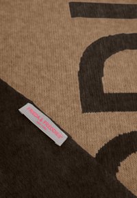 Bruine gebreide stof met grote zwarte letters; bevat een grijze label met "FRIEDA & FREDDIES" in het roze, gepositioneerd in de onderste hoek.
