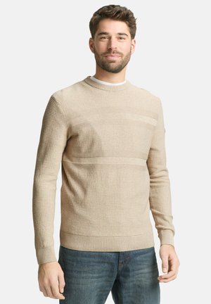 Homme portant un pull en tricot beige texturé avec des rayures horizontales et un jean bleu, debout devant un fond clair uni.