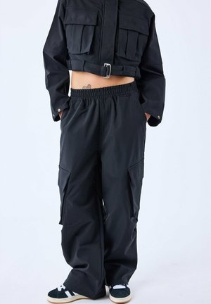 Chaqueta negra corta con dos bolsillos frontales y un cinturón; combinada con pantalones cargo negros sueltos que tienen bolsillos laterales y cinturilla elástica.