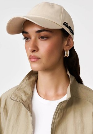 Młoda kobieta w beżowej czapce, kolczykach kołach, beżowej kurtce na zamek na białej koszuli, patrzy na bok z neutralnym wyrazem twarzy.