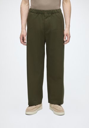 Persoană care poartă pantaloni largi verde măsliniu cu talie elastică și pantofi casual bej, stând pe un fundal deschis și simplu.